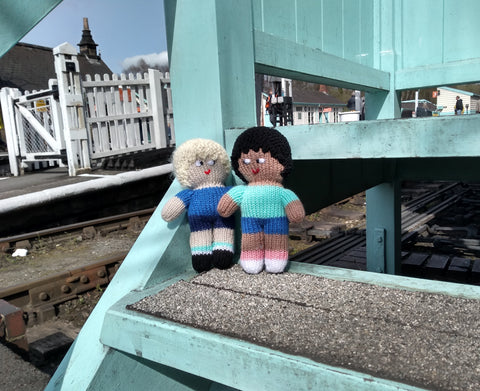 Two knitted Step Up For Nystagmus mascots tand on open steps at a railway station. They are wearing woolly trainers, shorts and T-shirts.